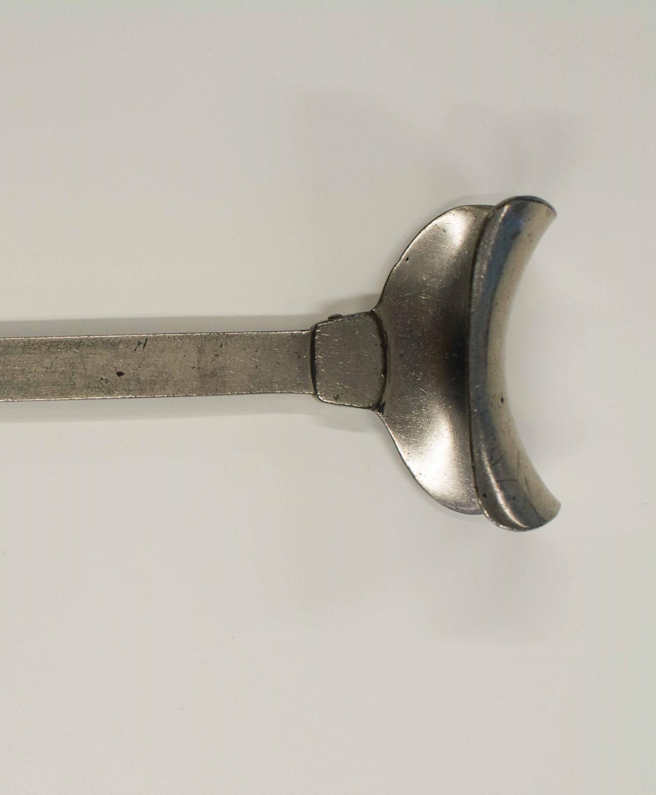 Instrumento metálico con forma de pala. Un extremo presenta forma de ancla, rematado con un aro y el extremo cuenta con una estructura cóncava