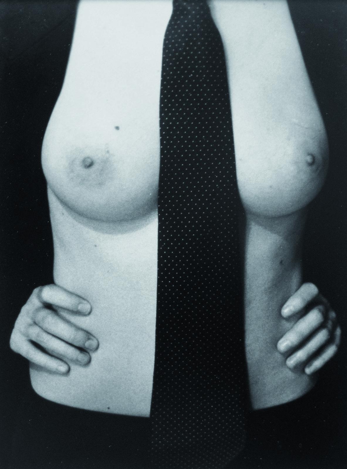 Fotografía en blanco y negro de un torso femenino desnudo. Entre los pechos cae una corbata negra. La mujer, cuyo rostro queda fuera de plano, viste chaqueta negra y ajusta las manos a su cintura