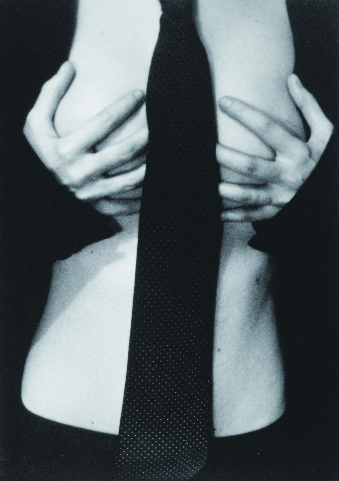 Fotografía en blanco y negro de un torso femenino desnudo. Entre los pechos, cubiertos por las manos, cae una corbata negra. La mujer, cuyo rostro queda fuera de plano, viste chaqueta negra 