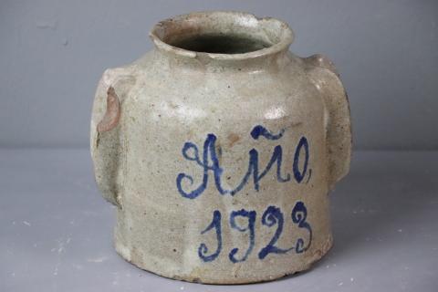 Tarro de cerámica de boca estrecha, asas con forma de herradura pegadas en los laterales. Esmalte blanco con inscripción en azul "Año 1923"